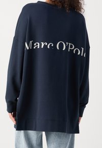 Sudadera oversize de color azul marino hecha de un tejido suave, con un logo impreso en blanco de forma horizontal, hombros caídos y puños de canalé.