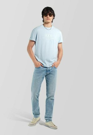 Junger Mann mit Sonnenbrille, hellblauem T-Shirt mit Grafik, hellblauer Jeans, beigen Sneakern, steht mit den Händen in den Taschen vor schlichtem Hintergrund.