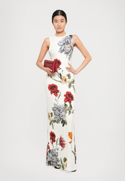 Woman standing with hand on hip wearing long white sleeveless dress with large colorful floral prints, holding red clutch bag.