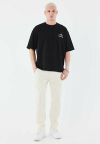 Camiseta de algodón negra de manga corta y cuello redondo, con un logo blanco en el lado izquierdo del pecho, combinada con pantalones blancos y zapatillas.