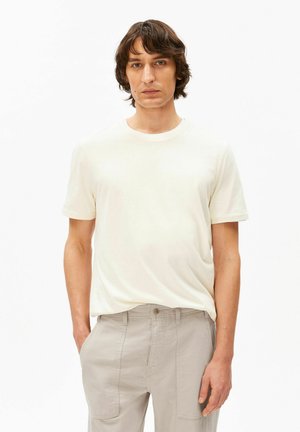 Jeune homme aux cheveux bruns portant un t-shirt à manches courtes uni blanc cassé et un pantalon beige clair, debout devant un fond blanc.