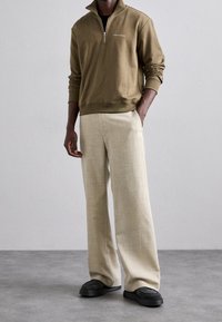 Felpa verde oliva con zip e colletto alto, abbinata a pantaloni beige a gamba larga e sneakers nere. Tessuto testurizzato e vestibilità comoda.