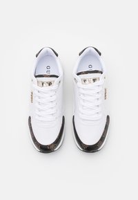 Paire de baskets blanches avec des accents à motifs noirs et un logo métallique "GUESS" sur la languette et le côté, vues de dessus sur un fond blanc.