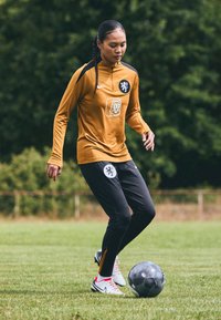 Nike sportsklær i oransje og svart, med et løveemblem. Spilleren dribler en svart fotball på grønt gress.