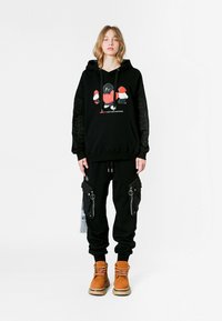 Joven con sudadera negra con gráfico en rojo y blanco y pantalones cargo negros con grandes bolsillos con cremallera, de pie contra un fondo blanco.