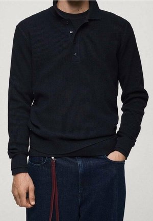 Mand iført en sort langærmet ribstrikket poloshirt og mørkeblå jeans med en vinrød nøglering, der hænger i bæltestroppen.