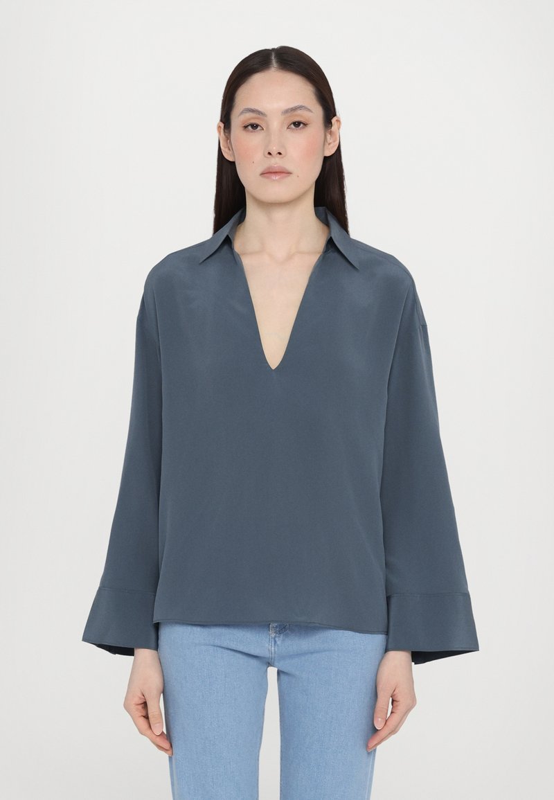 Blusa grigia scura con scollatura a V profonda, maniche lunghe a campana, realizzata in un tessuto morbido. Abbinata a jeans azzurri chiari. Design semplice e moderno.
