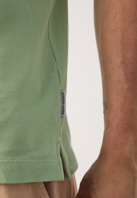 Grünes, strukturiertes Poloshirt mit Seitenschlitzen, versehen mit einem kontrastierenden Etikett, auf dem "SCOTCH & SODA" steht. Nahaufnahme des Stoffes und der Nähte.