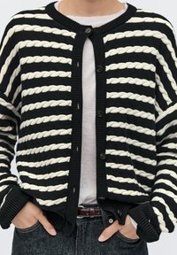 Černý a krémový pruhovaný kardigan s pletenou strukturou, s obsahujícími zapletené vzory, kulatým výstřihem a zapínáním na knoflíky. Nosit s bílou košilí.