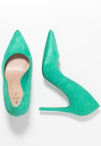 Talons hauts en daim bleu-vert avec un bout pointu, des côtés découpés et un talon stiletto fin. Texture lisse et design minimaliste.