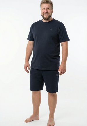 Smilende mand, stående barfodet iført matchende mørk navy T-shirt og shorts mod en ensfarvet hvid baggrund.