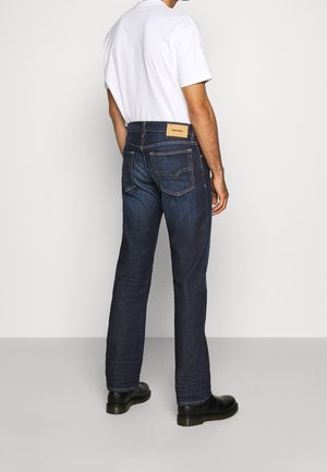 Hombre vistiendo vaqueros azul oscuro y zapatos negros, de pie de espaldas, con una camiseta blanca y una etiqueta de marca visible en la cintura.