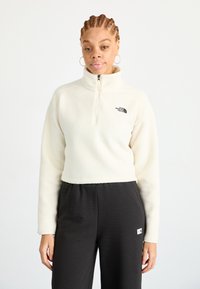 Pullover bianco in pile con zip fino a un quarto e logo nero sul lato sinistro del petto. Abbinato a pantaloni scuri e larghi con un piccolo etichetta del logo.