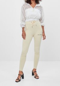 Béžové slim-fit tepláky s elastickým pasem a šňůrkou. Doplněné bílou průhlednou zkrácenou bundou a černými sandály na podpatku.