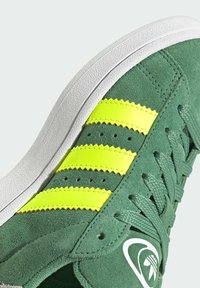 Zapatillas de ante verde con tres franjas de neón amarillas, suela de goma blanca y detalles texturizados. Incluye cordones tejidos y perforaciones.