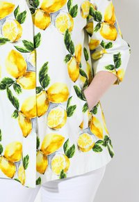 Blusa bianca con motivo di limoni e foglie, caratterizzata da limoni gialli vibranti e foglie verdi, maniche a tre quarti e tasche laterali.