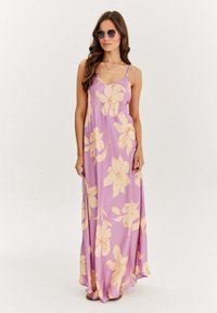 Maxi šaty z fialové látky s velkými krémovými květinovými vzory, tenkými ramínky a splývavým designem. Nosené s slunečními brýlemi, dlouhé vlasy jemně padají.
