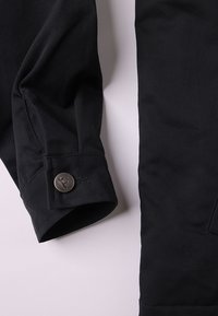 Μανίκι μαύρου υφασμάτινου σακακιού με κουμπί που φέρει ανάγλυφο λογότυπο, παρουσιάζοντας λεία υφή και ραμμένη εφαρμογή.