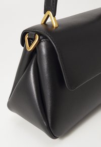 Borsa a tracolla in pelle nera con una forma strutturata, dotata di una patta pieghevole e dettagli hardware triangolari in oro. Texture liscia, design minimale.