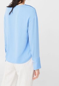 Lyseblå skjorte med lange ærmer og rundet kant, knapdetaljer på ærmerne og afslappet pasform. Stribede bukser er delvist synlige.