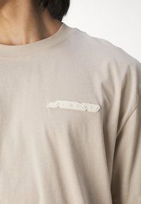 Beige bomulds T-shirt med et hævet hvidt logo på venstre bryst, rund halsudskæring og korte ærmer, glat tekstur med subtil stræk.