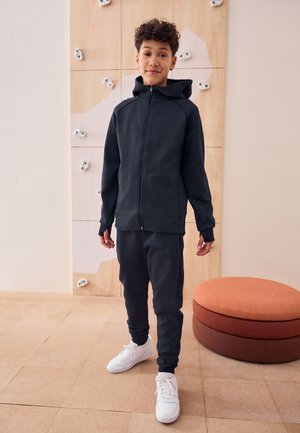 Jongen met krullend haar die een zwarte trui met rits, zwarte broek en witte sneakers draagt, staat binnen in de buurt van een klimwand en een rond kussen.