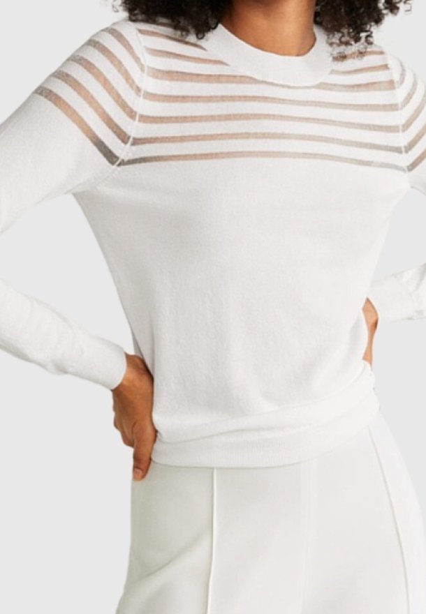 Femme portant un pull blanc avec des rayures horizontales beige sur le haut de la poitrine et les épaules, associé à un pantalon blanc.