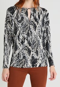 Koszula w czarno-białe wzory węża z dekoltem w kształcie dziurki, długimi rękawami i luźnym krojem. Miękki materiał z delikatnym połyskiem. Połączona z brązowymi spodniami.