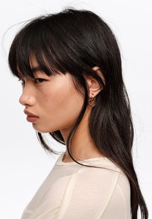 Młoda kobieta o długich czarnych włosach z grzywką, ubrana w kremową bluzkę i złote kolczyki koła, zwrócona w lewo na białym tle.