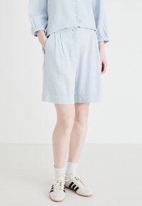 Pantaloni corti blu chiaro in misto lino con pieghe, tasche laterali e bottone frontale. Abbinati a calzini bianchi e sneakers a strisce nere.