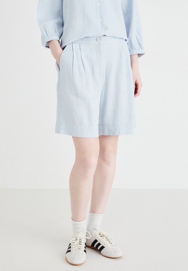 Pantaloni corti blu chiaro in misto lino con pieghe, tasche laterali e bottone frontale. Abbinati a calzini bianchi e sneakers a strisce nere.
