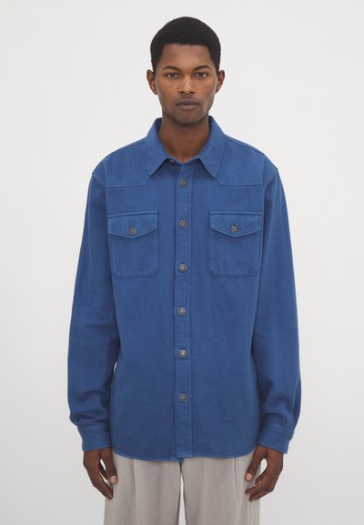 Camicia di cotone blu con chiusura a bottoni, due tasche sul petto, colletto strutturato e maniche lunghe. Tessuto liscio con bottoni argentati.