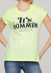 Chemise à manches courtes jaune fluo avec capuche, ornée d'un texte en noir "C'est l'été SOCCX" et de strass décoratifs. Tissu doux et léger.