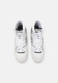 Pár bílých vysoce chodících tenisek New Balance 650 se šněrováním, černými logovými akcenty a polstrovanými límci na bílém pozadí.