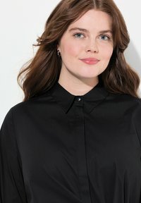 Camisa negra de botones con cuello puntiagudo, hecha de una tela suave. Presenta botones en la parte delantera y una silueta ajustada y elegante.