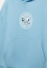 Lyseblå sweatshirt med "Peanuts Hockey Club" grafik, hvor Snoopy holder en hockeykølle. Indeholder en frontlomme og har en blød tekstur.