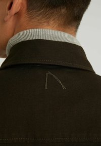 Brązowa kurtka z tkaniny z szarym żeberkowanym kołnierzem i haftowanym wzorem w jaśniejszej nici na plecach. Zbliżenie na obszar kołnierza.