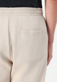 Beige nadrág rugalmas derékrésszel és egy hátsó zsebbel, lekerekített sarkakkal és látható varrási részletekkel. Simább textúrájú.