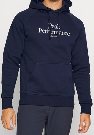 Mann trägt einen dunkelblauen Hoodie mit Fronttasche und dem Text "Peak Performance Est. 1986" auf der Brust.