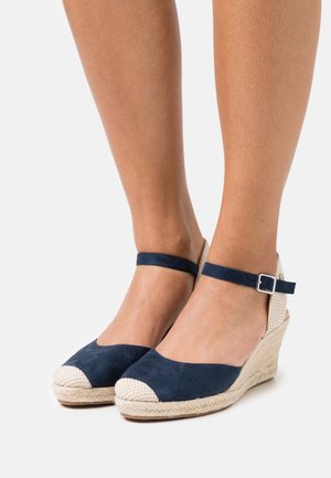 Sandales compensées en suède navy avec un talon en jute tissé et un bout beige contrastant. La lanière de cheville est dotée d'une boucle.