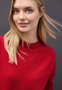 Maglione rosso con scollo a girocollo a coste, texture morbida e design con spalle basse, con capelli biondi che incorniciano uno sfondo neutro.