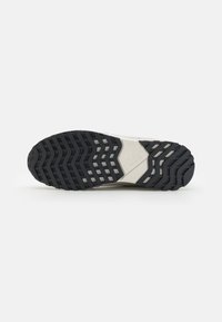 Semelle de sneaker blanche avec un motif de traction en caoutchouc noir, présentant une surface texturée et des rainures prononcées pour une meilleure adhérence.