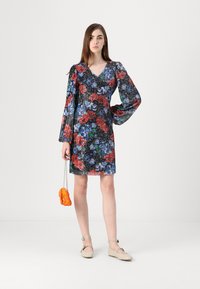 Robe florale avec de longues manches bouffantes, fond noir, roses colorées en rouge et bleu. Associée à un sac à main orange en peluche et des chaussures beiges.