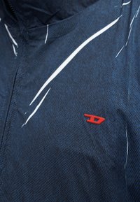 Σκούρο μπλε υφασμάτινο μπουφάν με μικρό κόκκινο λογότυπο στο στήθος και γυαλιστερή αντανακλαστική επιφάνεια στο επάνω αριστερό μέρος.