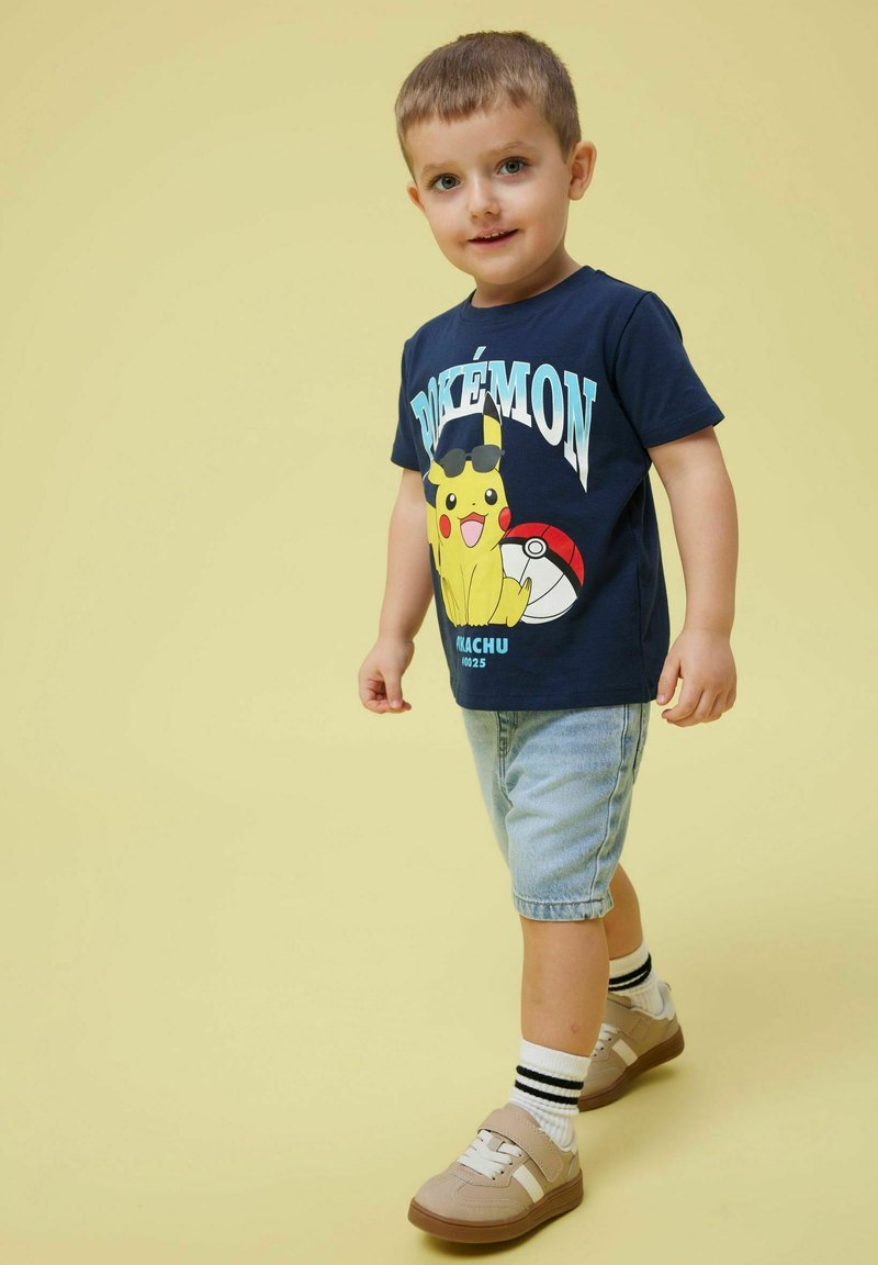 Camiseta de Pokémon azul marino con Pikachu usando gafas de sol, combinada con pantalones cortos de mezclilla clara y zapatillas beige con calcetines a rayas en blanco y negro.