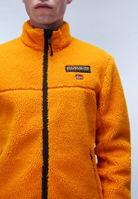 Fleece jacket in bright orange with a high collar, black zipper, and logo patch featuring a Norwegian flag. Textured, soft fabric.