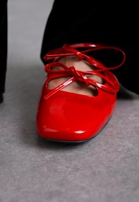 Red patent leather shoe with a pointed toe and crisscross lace-up design. Smooth texture and polished finish. Non-slip black sole.