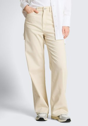 Personne portant un pantalon beige à jambes larges, une chemise blanche à boutons, et des baskets blanc-gris, debout avec une main dans la poche.