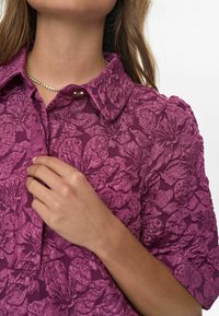 Cămașă cu model floral purpuriu, dintr-un material texturat. Prezintă un guler clasic și mâneci scurte, cu un detaliu de tip placket cu nasturi.