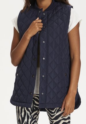 Persoon draagt een navy blauwe gewatteerde bodywarmer zonder mouwen over een wit shirt met korte mouwen en zebra-print broek, houdt met één hand de kraag van de bodywarmer vast.